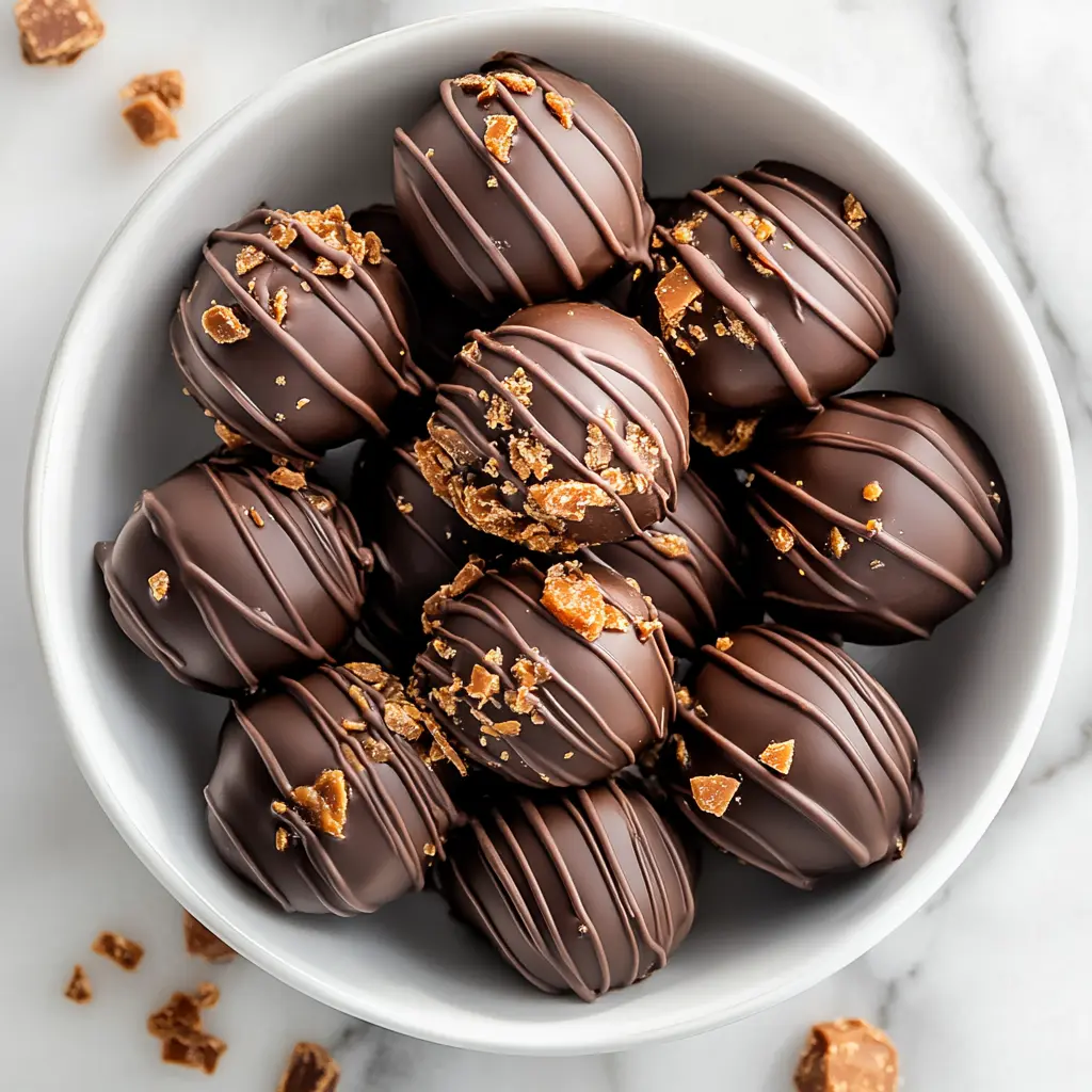 Featured final dish of Irresistible Butterfinger Balls in a white porcelain bowl with chocolate drizzle on marble.